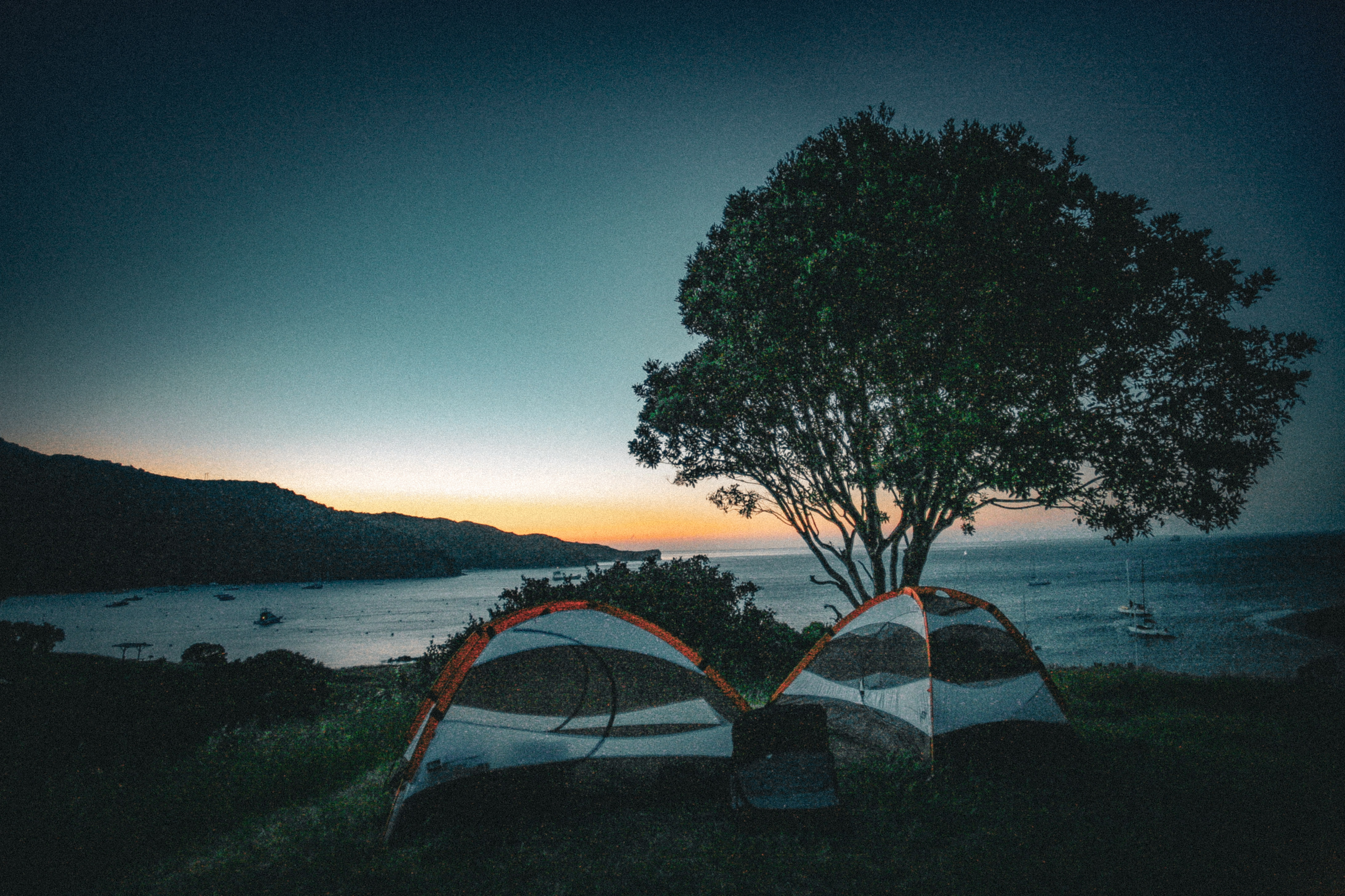 Two camping tents on a grassy bluff at twilight overlooking a calm bay with anchored boats