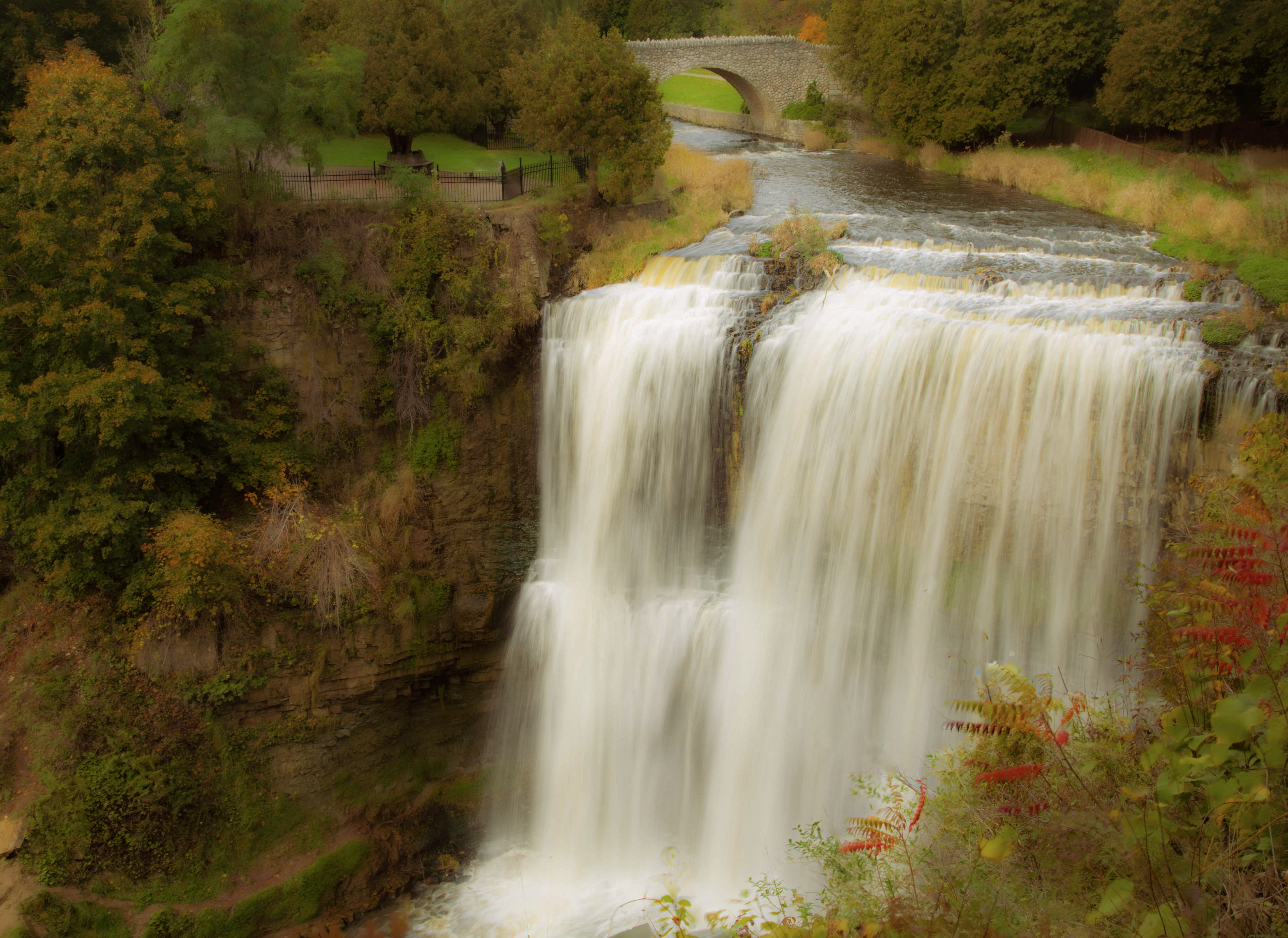 Wide autumn waterfall cascading over rocky tiers toward a stone arch bridge in a wooded gorge