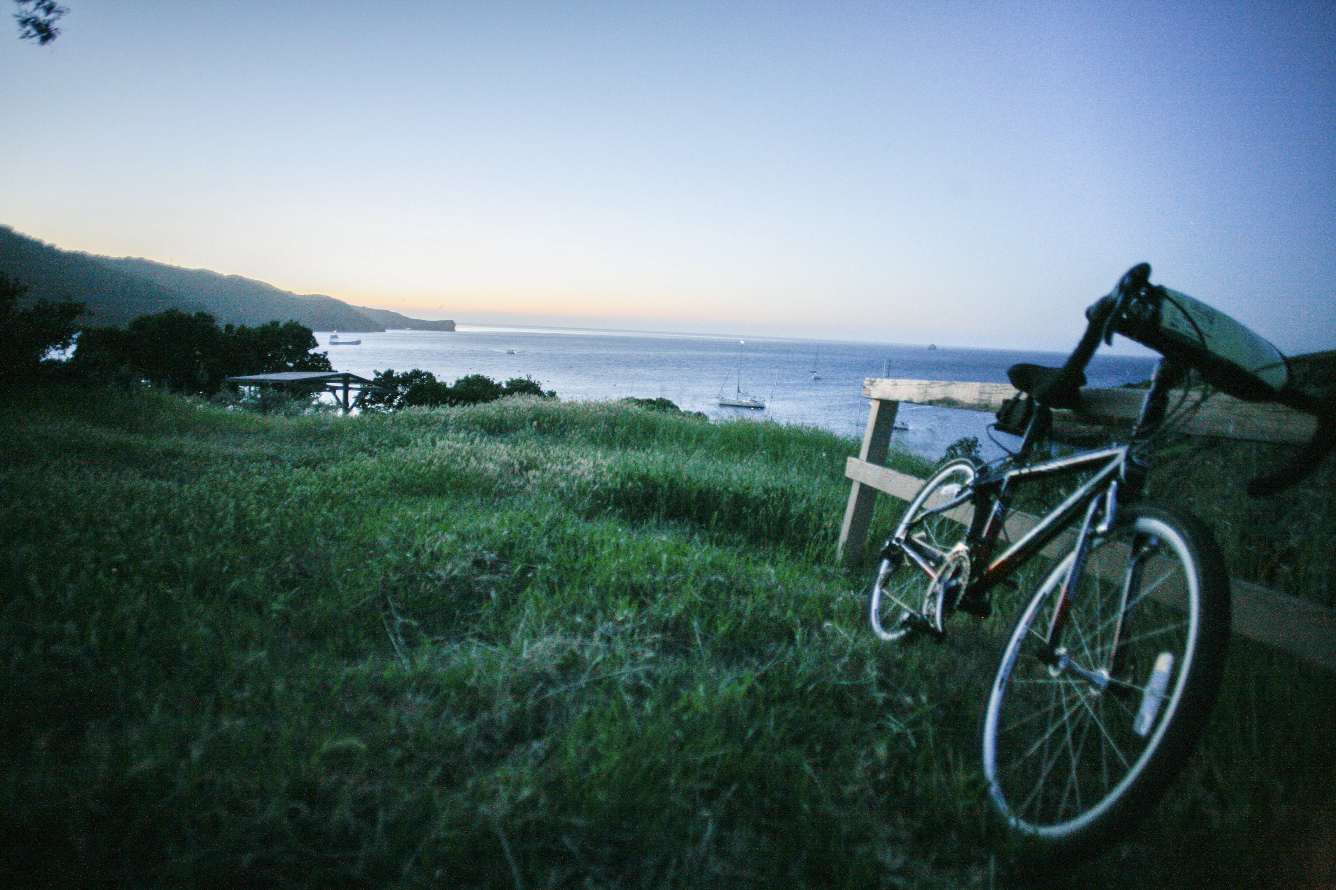 A touring bicycle leans against a wooden fence on a grassy hill overlooking a quiet bay at twilight