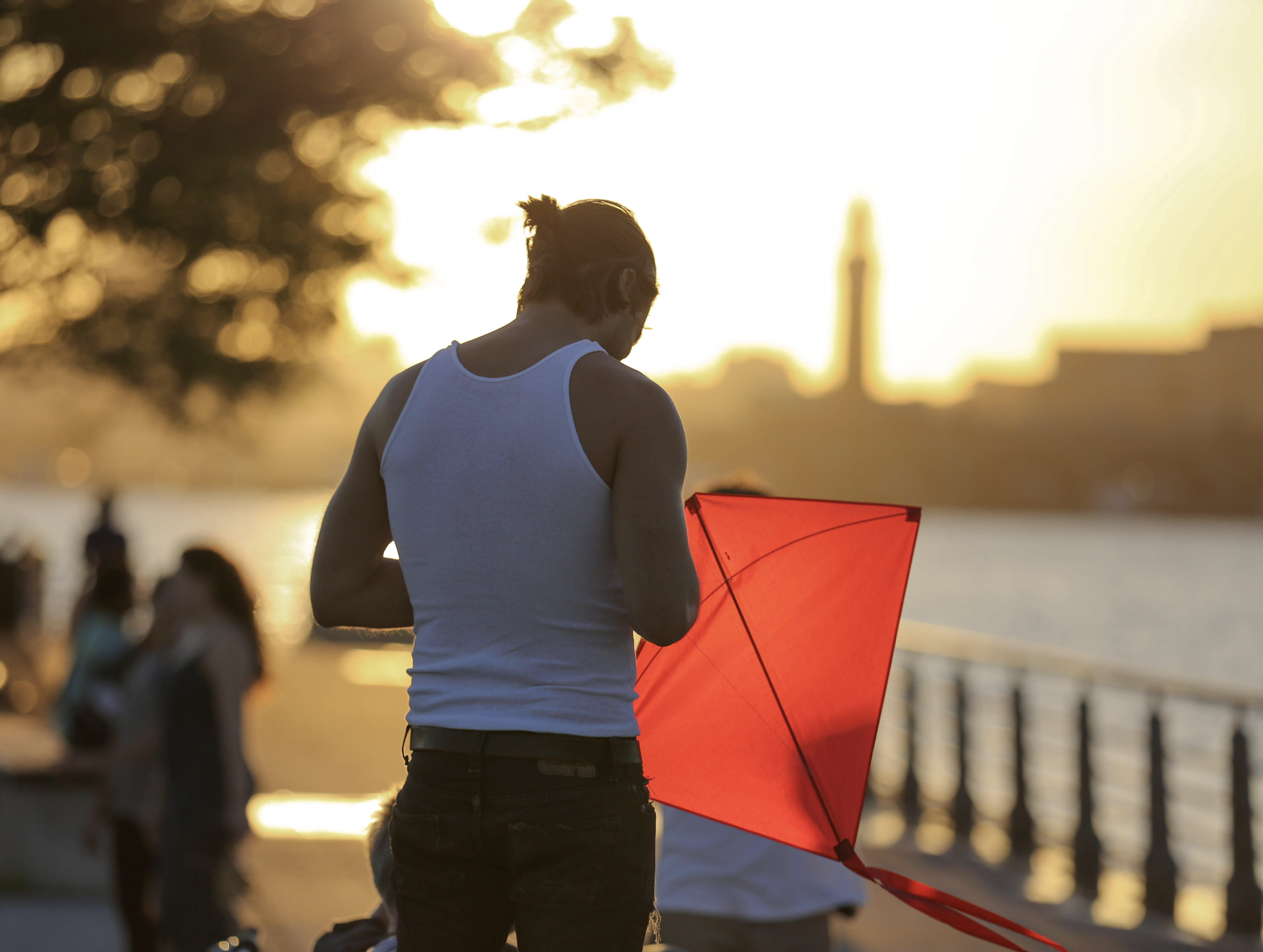 A man adjusts a bright red kite on a waterfront promenade at golden hour with a distant city skyline across the water