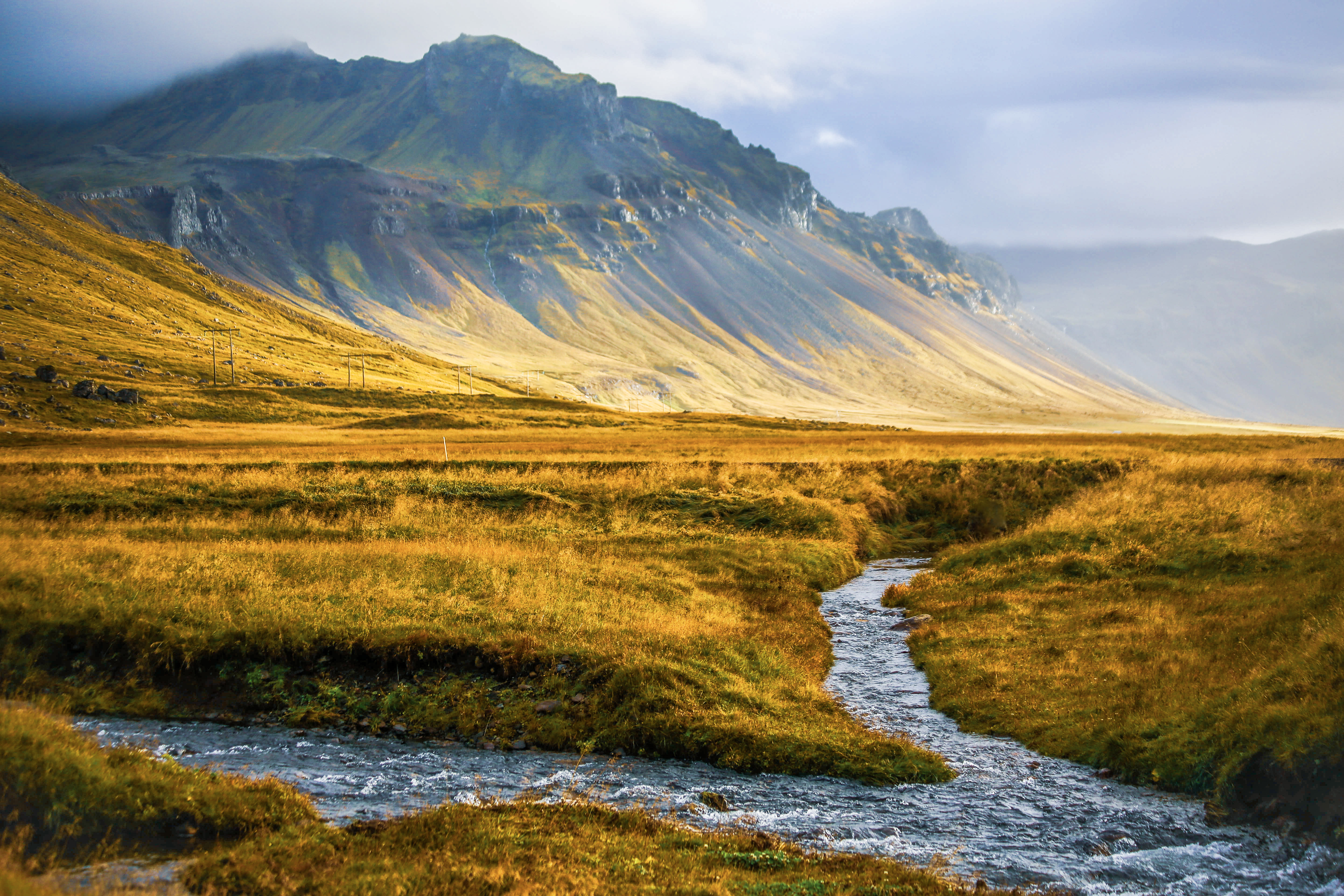 Golden grassy plain and clear stream beneath steep mossy mountains under low clouds