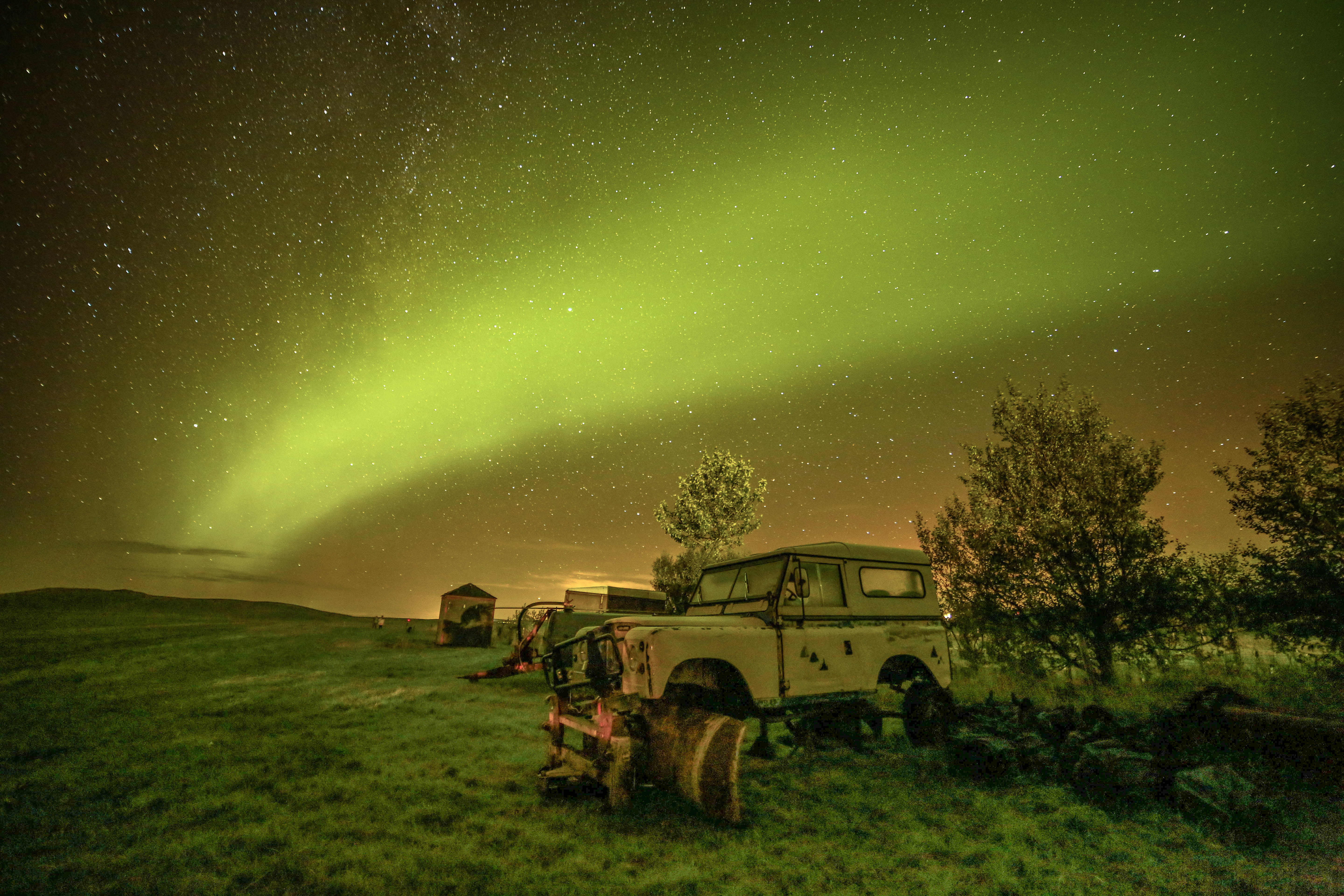 Abandoned vehicles in a grassy field under a green aurora and a starry sky, with a small hut and trees at the edge of the field.