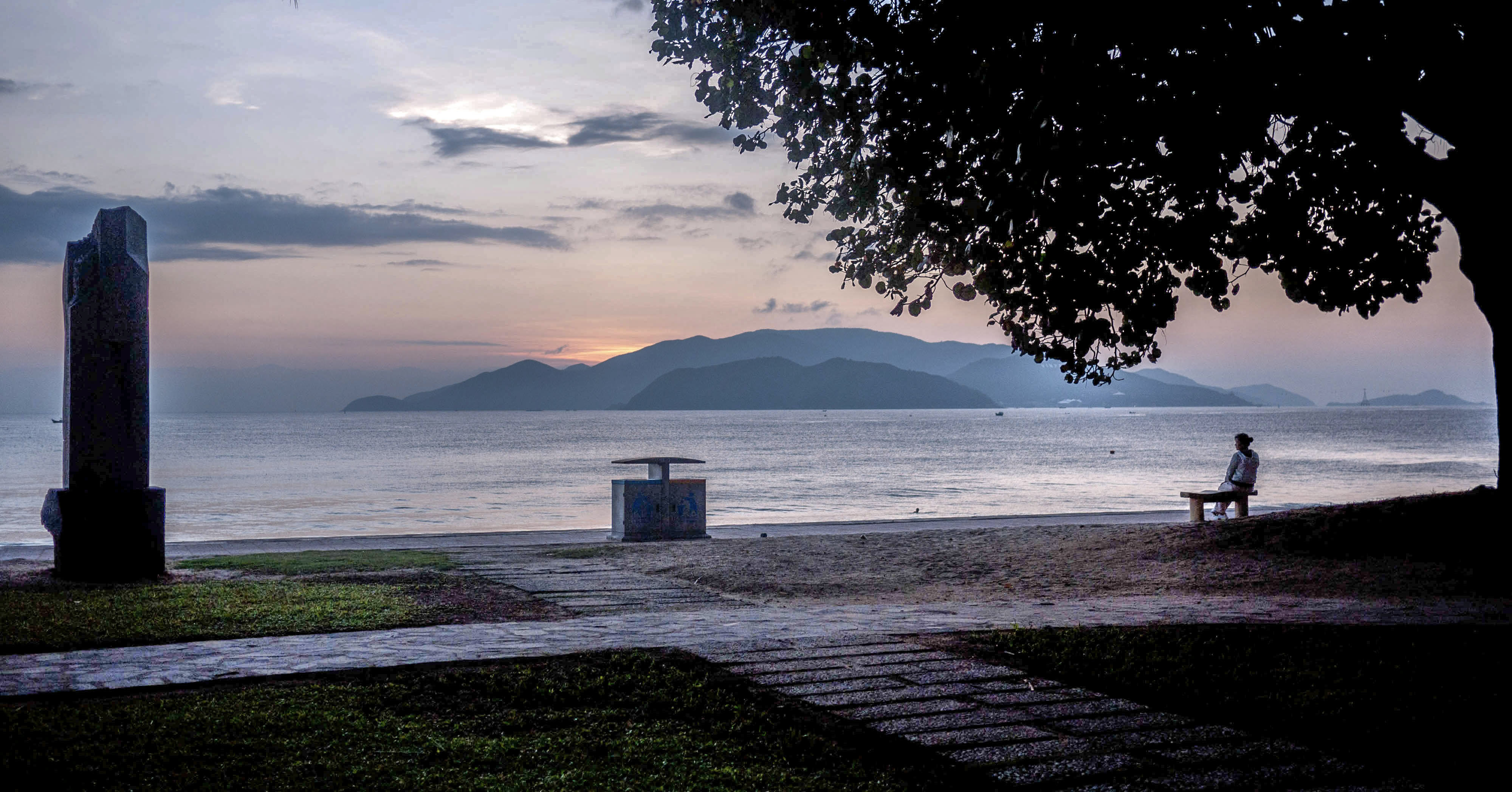 Silhouetted figure on a shoreline bench at dusk facing calm water and distant mountains