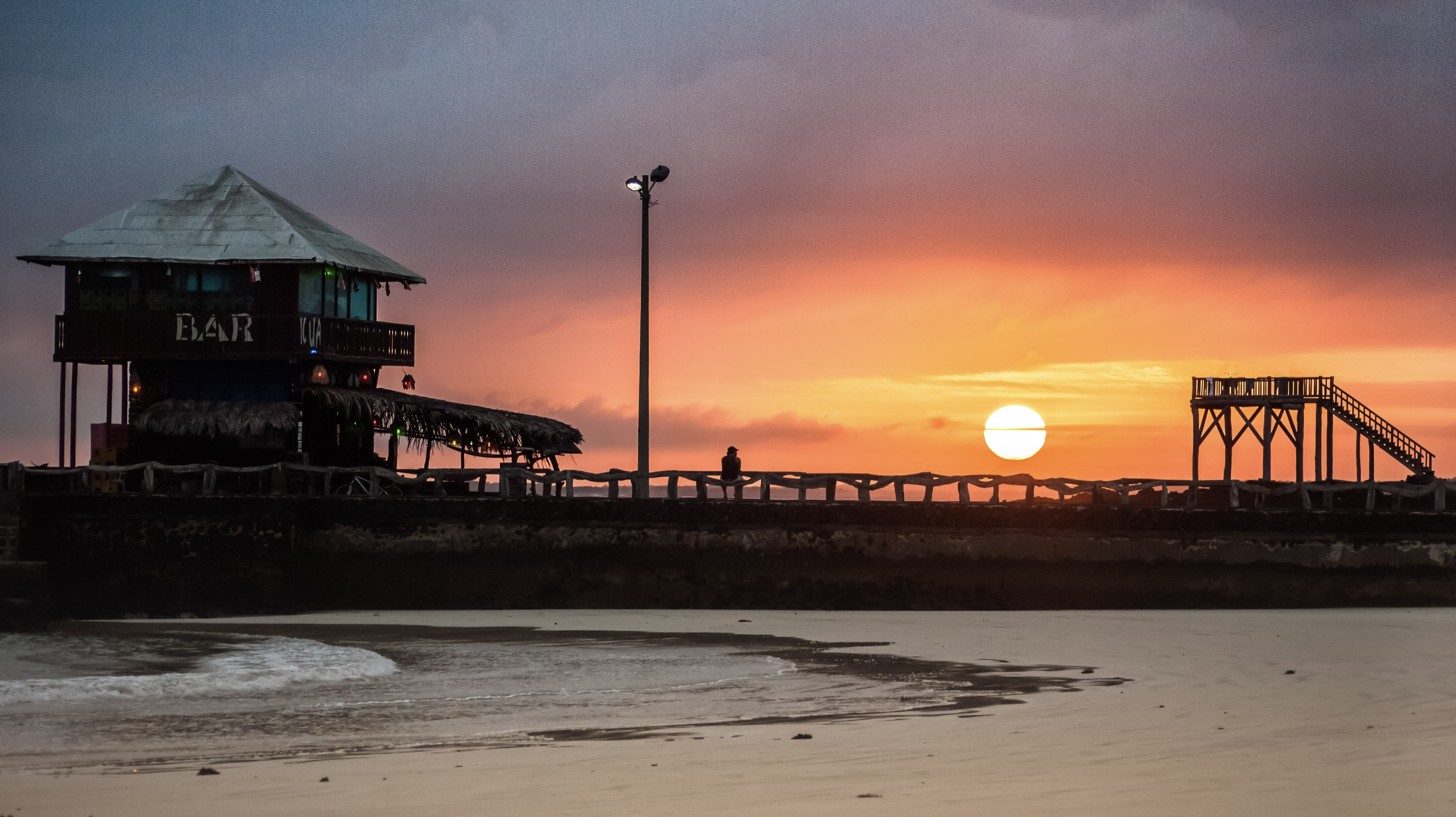 Tropical beach pier at sunset with a wooden bar building silhouetted against an orange sky.